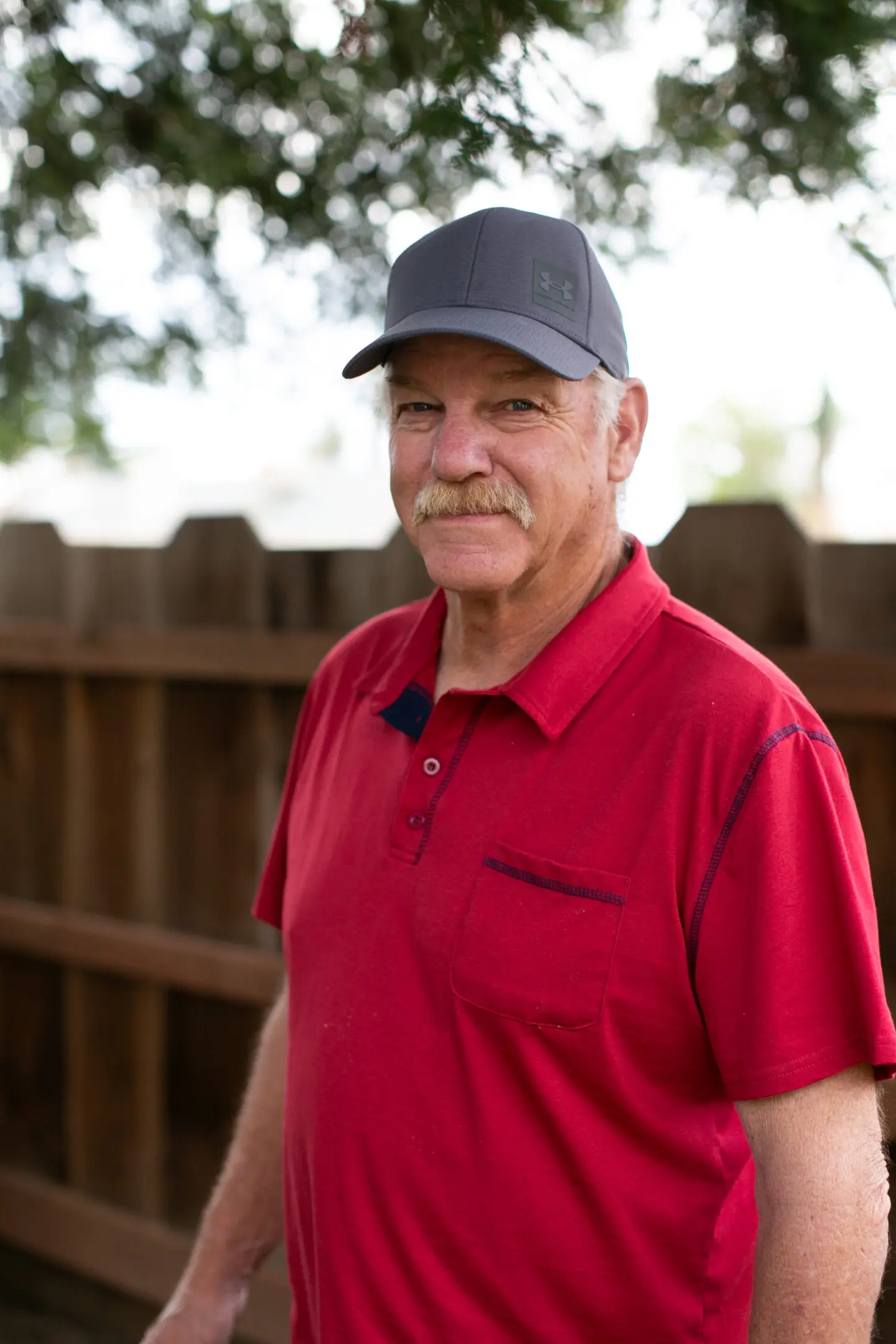 Mike Stevens smiling, wearing a Maroon Polo and ball cap, standing in front of a wood fence